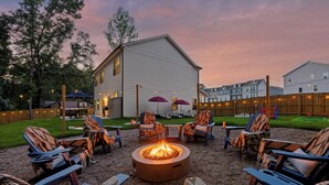 An outdoor seating area with a fire pit surrounded by lounge chairs, a wooden fence, and a building in the background. - New! Sleeps 20 — Hot Tub, Minigolf, Firepit,Outdoor Movie & Game Room (Charlotte)
