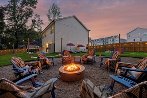 An outdoor seating area with a fire pit surrounded by lounge chairs, a wooden fence, and a building in the background. - New! Sleeps 20 — Hot Tub, Minigolf, Firepit,Outdoor Movie & Game Room (Charlotte)