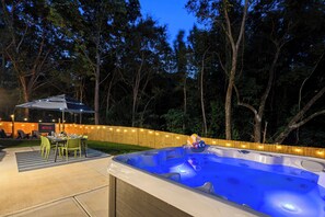A backyard with a hot tub, a dining table with chairs, and a wooden fence with string lights.