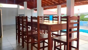 A dining area with a wooden table and chairs, a pool in the background, and a covered patio with a wooden ceiling. - SHB Chácara para Eventos com Piscina (Elias Fausto)