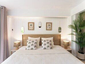 A bedroom with a wooden headboard, two bedside tables with lamps, a large potted plant, and wall-mounted artwork. - Villa Belle infinity pool and unique charm featuring 3 air-conditioned bedrooms (Grand-Bourg)
