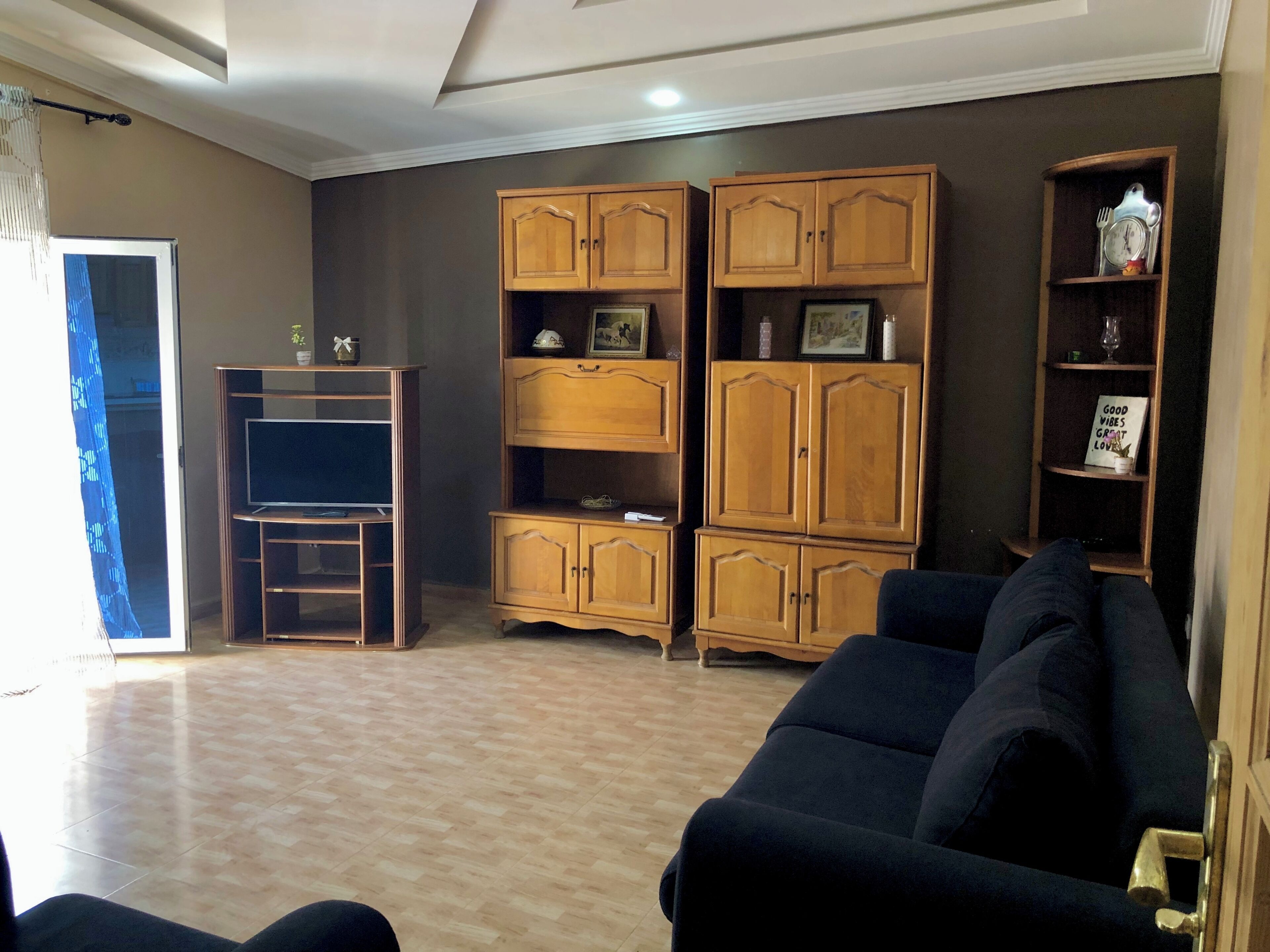 Uma sala de estar com um sofá escuro, armários de madeira, uma televisão sobre um móvel e uma pequena estante com objetos decorativos.