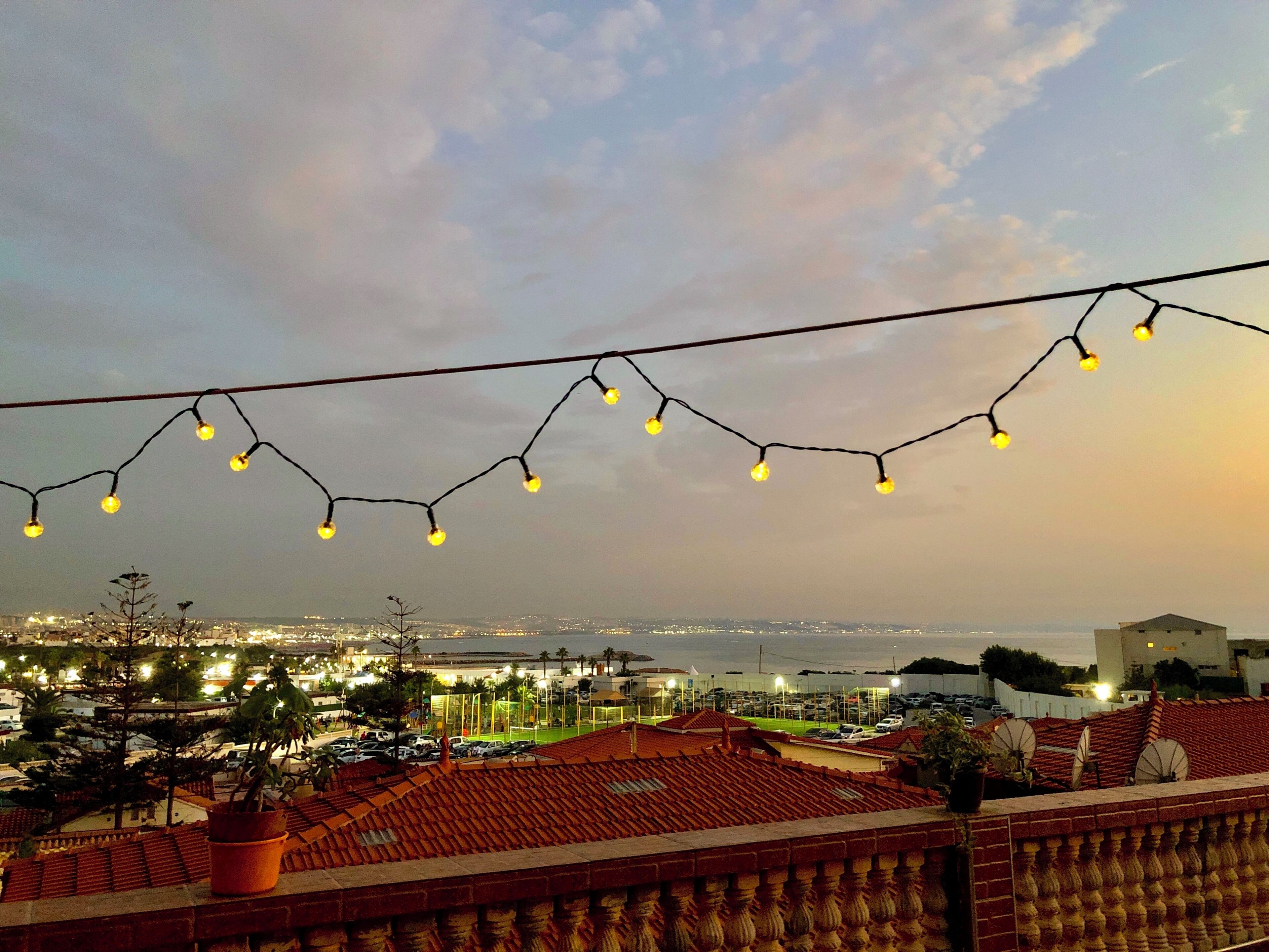 A balcony with a view of a cityscape at dusk, featuring a string of lights and a potted plant.