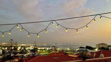 Ein Balkon mit Blick auf eine Stadtlandschaft im Abenddämmerung, mit Lichterkette und einer Pflanze im Topf.