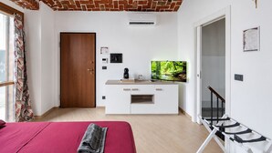 Ein Zimmer mit Ziegeldecke, einem Bett mit roter Bettdecke, einem Fernseher auf einem weißen Schrank und einem Fenster mit Vorhängen.