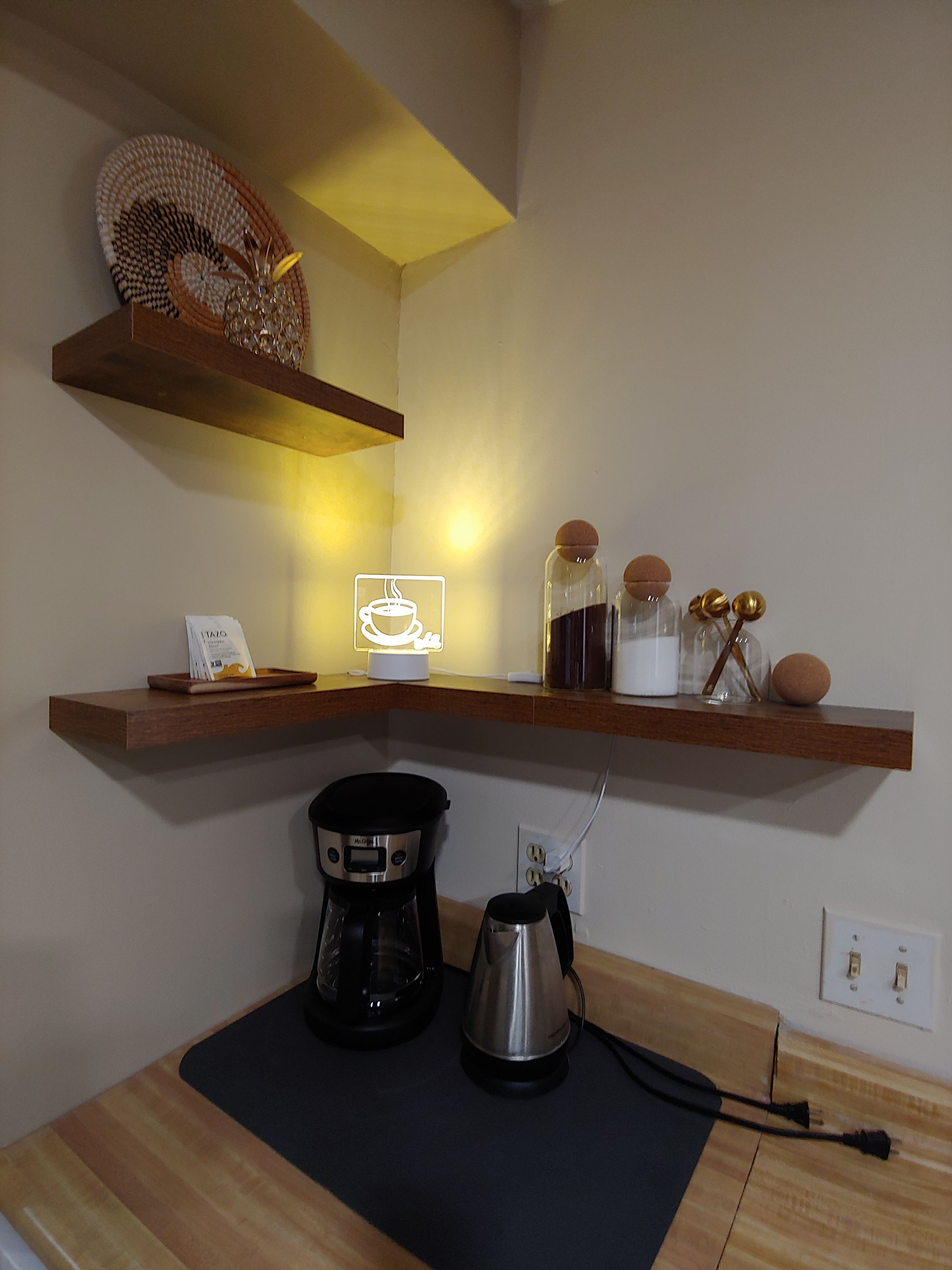 A kitchen with wooden shelves, a coffee maker, a kettle, and a sign indicating a coffee service.