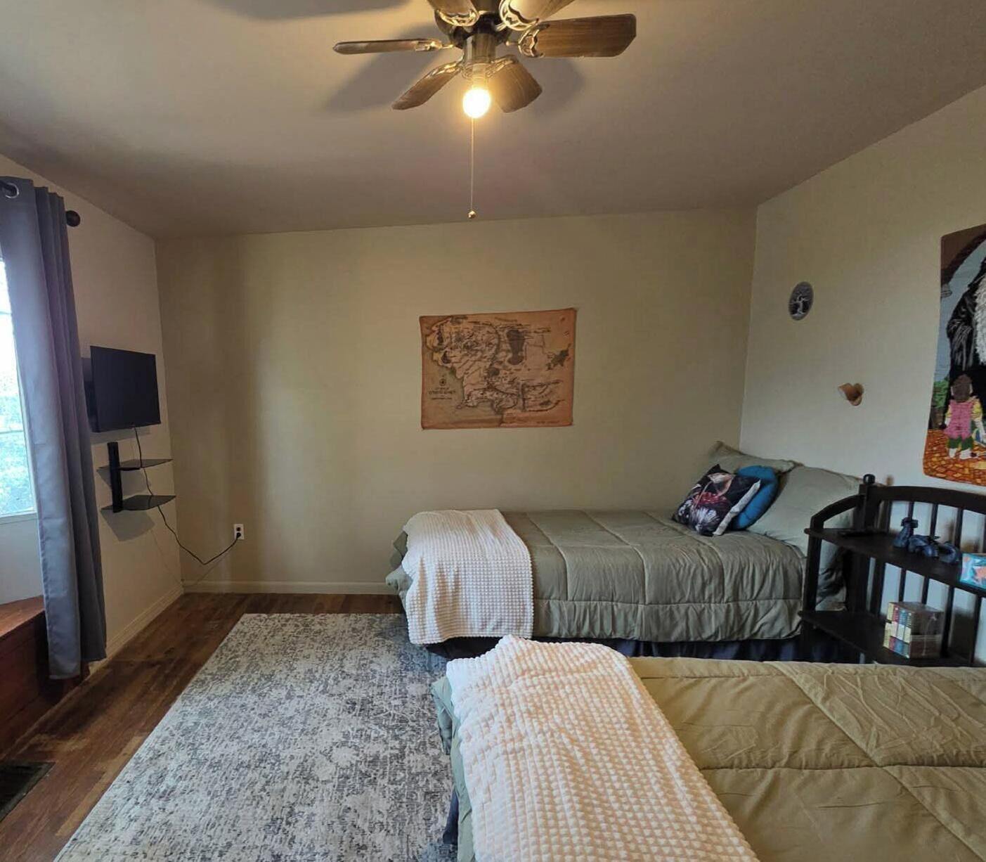 A bedroom with a bed, a desk, a chair, a ceiling fan, a map on the wall, and a window with curtains.