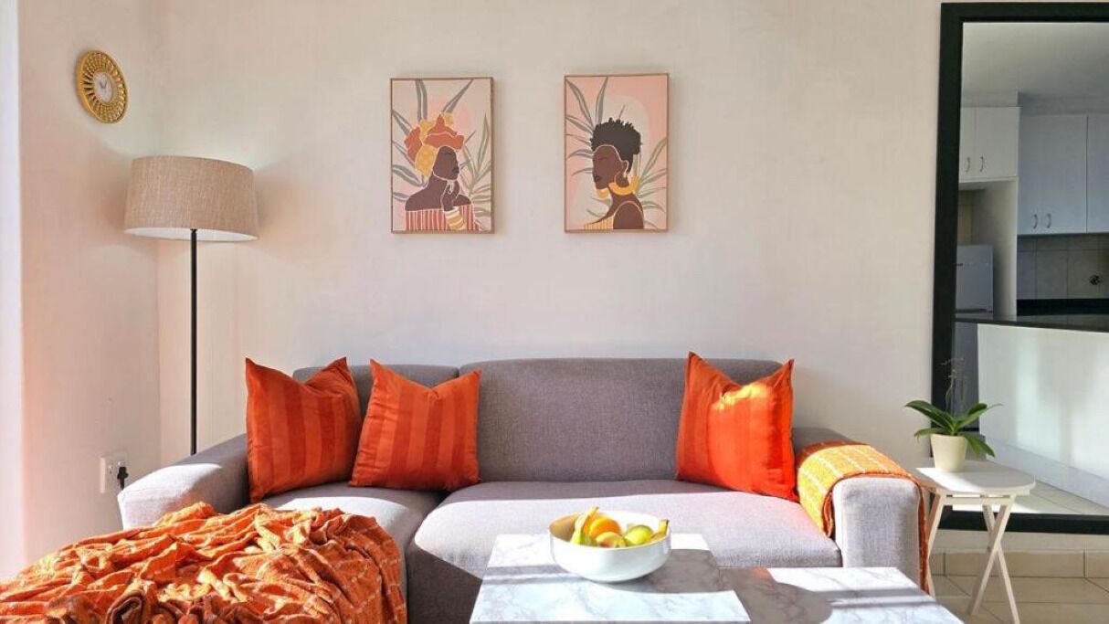 A modern living room with a grey sofa, orange cushions, a white coffee table with fruit, and two wall art pieces.