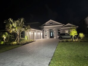 A two-story house with a paved driveway leading to a garage, surrounded by well-maintained landscaping and a parked car. - Newly built 4-bed house in Esplanade by the Islands BOAT INCLUDED with captain. (Naples)