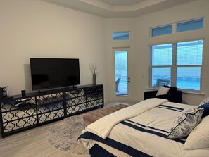 A bedroom with a bed, a TV on a black console, a door leading outside, and large windows.
