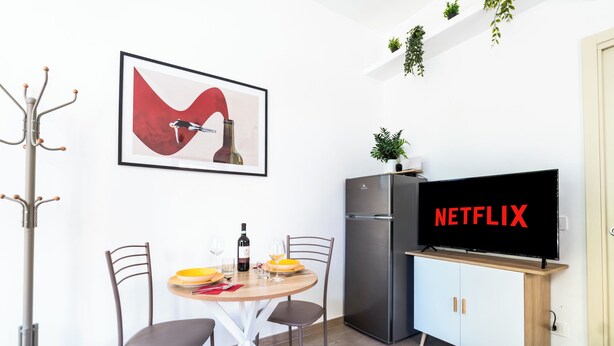 Eine moderne Küche mit Esstisch, Stühlen, einem Kühlschrank, einem Fernseher, der 'NETFLIX' zeigt, und einem gerahmten Kunstwerk an der Wand.