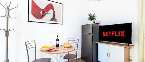A modern kitchen with a dining table, chairs, a refrigerator, a TV displaying 'NETFLIX,' and a framed artwork on the wall.