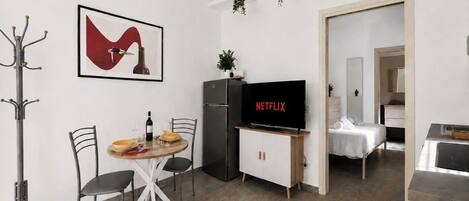 Un espacio habitable compacto con comedor, kitchenette, televisor mostrando 'NETFLIX' y una pequeña zona de estar.