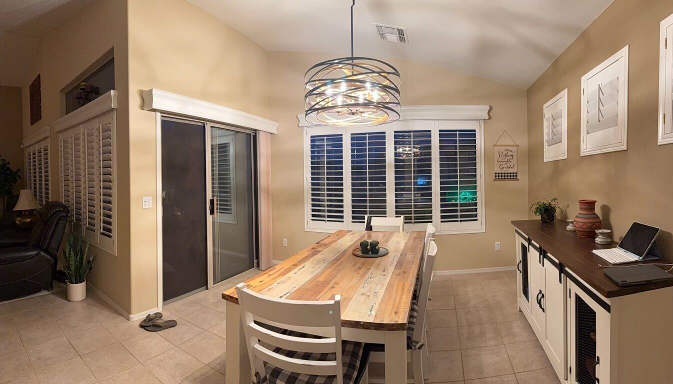 A dining area with a wooden table, chairs, a pendant light, a sliding glass door, and a kitchen area with cabinets and a laptop.