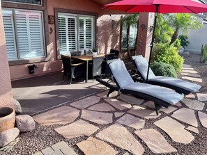 Un patio con suelo de piedra, una mesa con sillas y dos sillones de descanso bajo un paraguas rojo.
