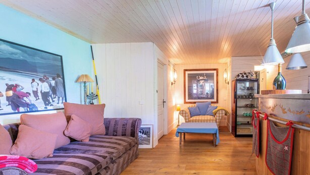 Ein gemütliches Wohnzimmer mit einer Couch, einem Tisch und einer Küchenzeile mit Kühlschrank und einem aufgehängten Topflappen.