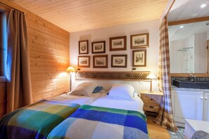 Une chambre comprenant un lit, deux tables de chevet, un mur orné de tableaux encadrés, une fenêtre avec des rideaux et une salle de bain visible à travers une porte en verre.