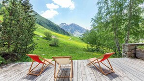 Eine Holzterrasse mit zwei roten Liegestühlen, die auf eine gebirgige Landschaft ausgerichtet ist.