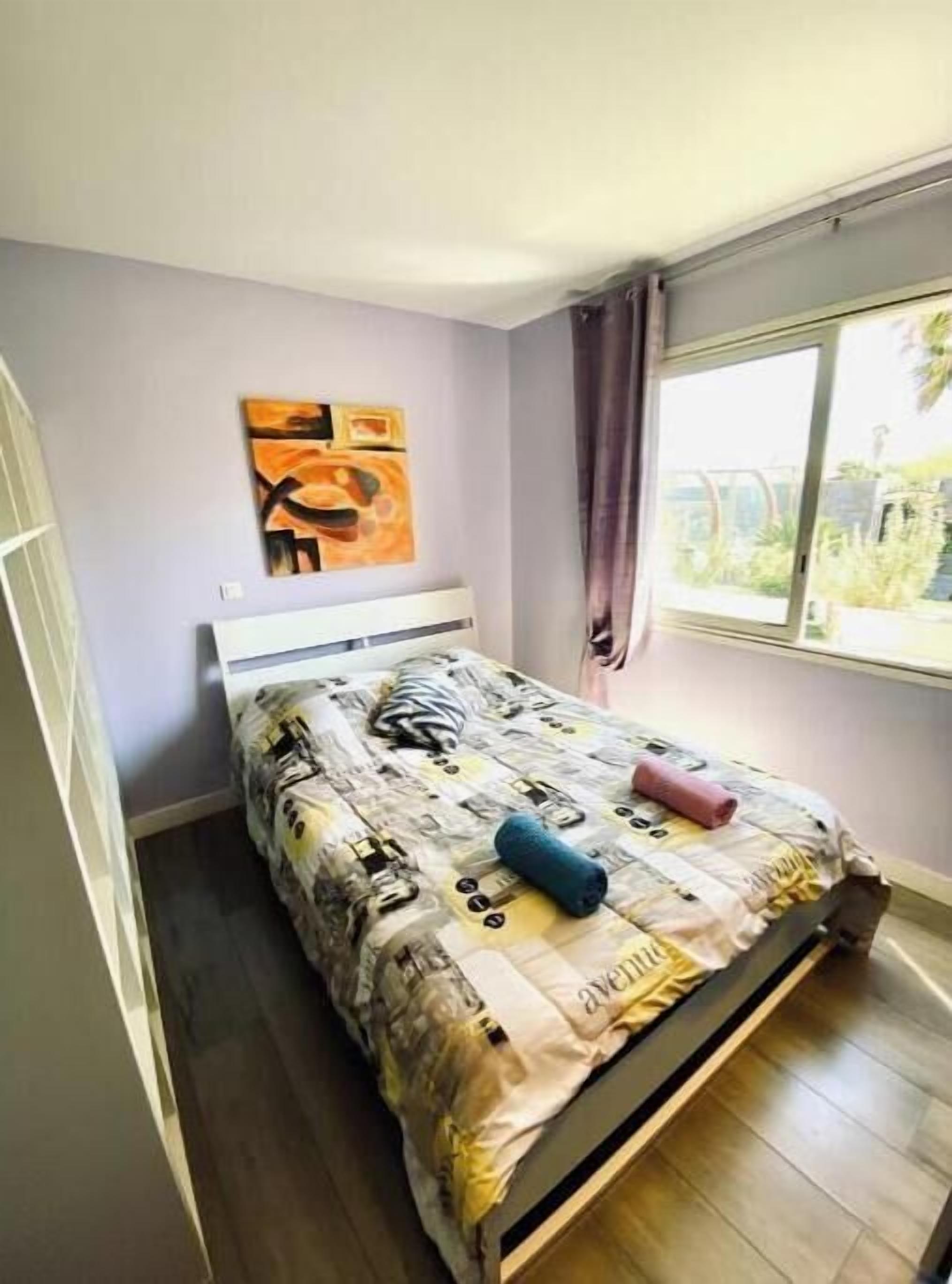 Un dormitorio con cama, ventana con cortinas, una obra de arte en la pared y suelo de madera.