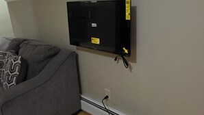 Ein Zimmer mit grauem Sofa, einem an der Wand befestigten Flachbildfernseher, einem Standventilator und einer Steckdose.