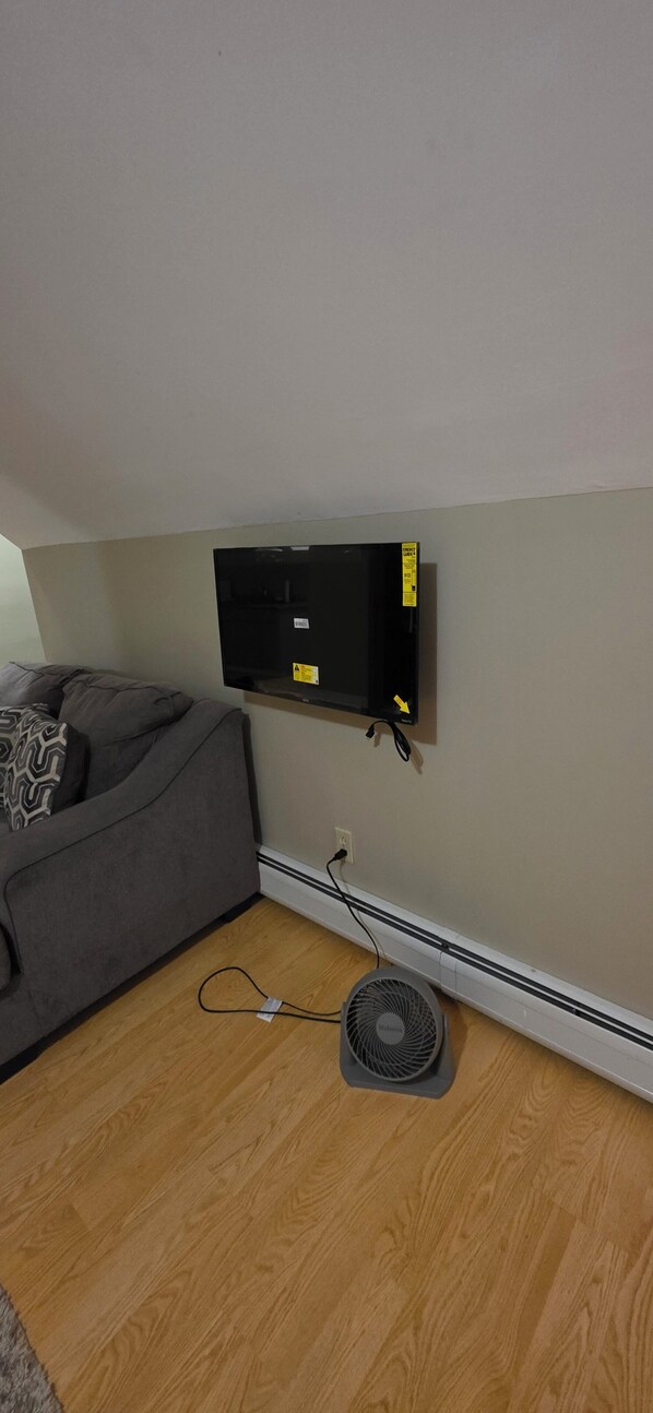 Una habitación con un sofá gris, un televisor de pantalla plana montado, un ventilador de pie y un enchufe en la pared.