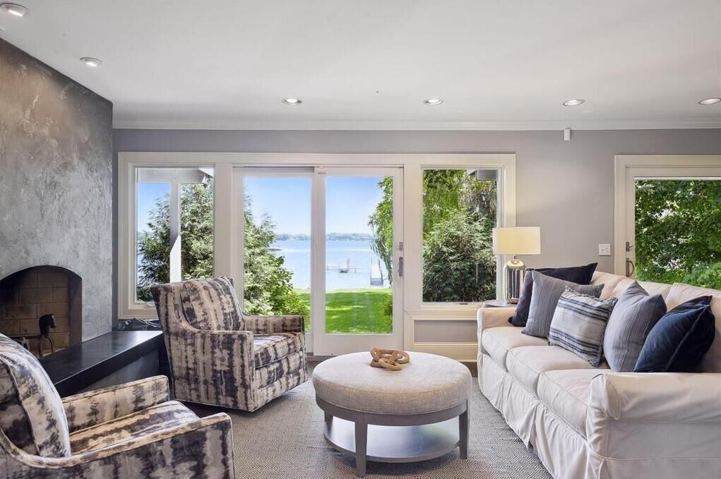 A living room with a fireplace, a sofa, a round ottoman, and a view of a body of water through large windows.