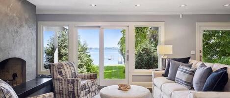 A living room with a fireplace, a sofa, a round ottoman, and a view of a body of water through large windows.