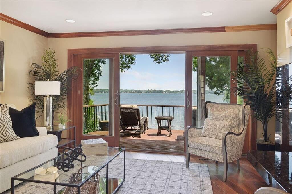 A living room with a sofa, coffee table, and armchairs, a glass coffee table, and a view of a lake through glass doors.