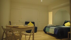 Ein Zimmer mit Esstisch, zwei Stühlen, einem Bett mit blaugrüner Bettdecke und einer weißen Tür.