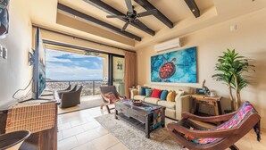 A spacious living area with a balcony, a ceiling fan, a sofa with colorful cushions, and a large painting of a turtle. - CCV - 2BR Ocean-View Condo w/ Balcony, Golf & Pools at Quivira - Cabo San Lucas (Cabo San Lucas)