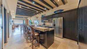 A modern kitchen with a central island, bar stools, stainless steel appliances, and a dining area with a view. - CCV - 2BR Ocean-View Condo w/ Balcony, Golf & Pools at Quivira - Cabo San Lucas (Cabo San Lucas)