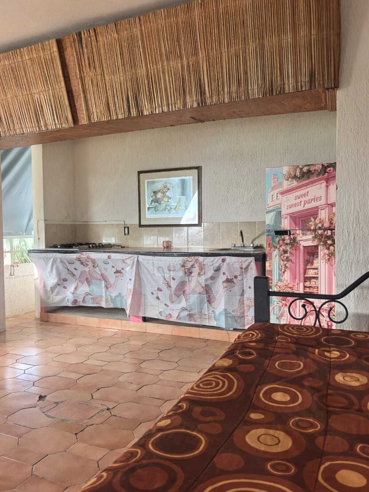 Eine Küchenzeile mit Fliesenboden, einem Tisch mit Blumenmuster-Tischdecke, einem gerahmten Bild an der Wand und einem Poster mit einer Bäckerei-Szene.