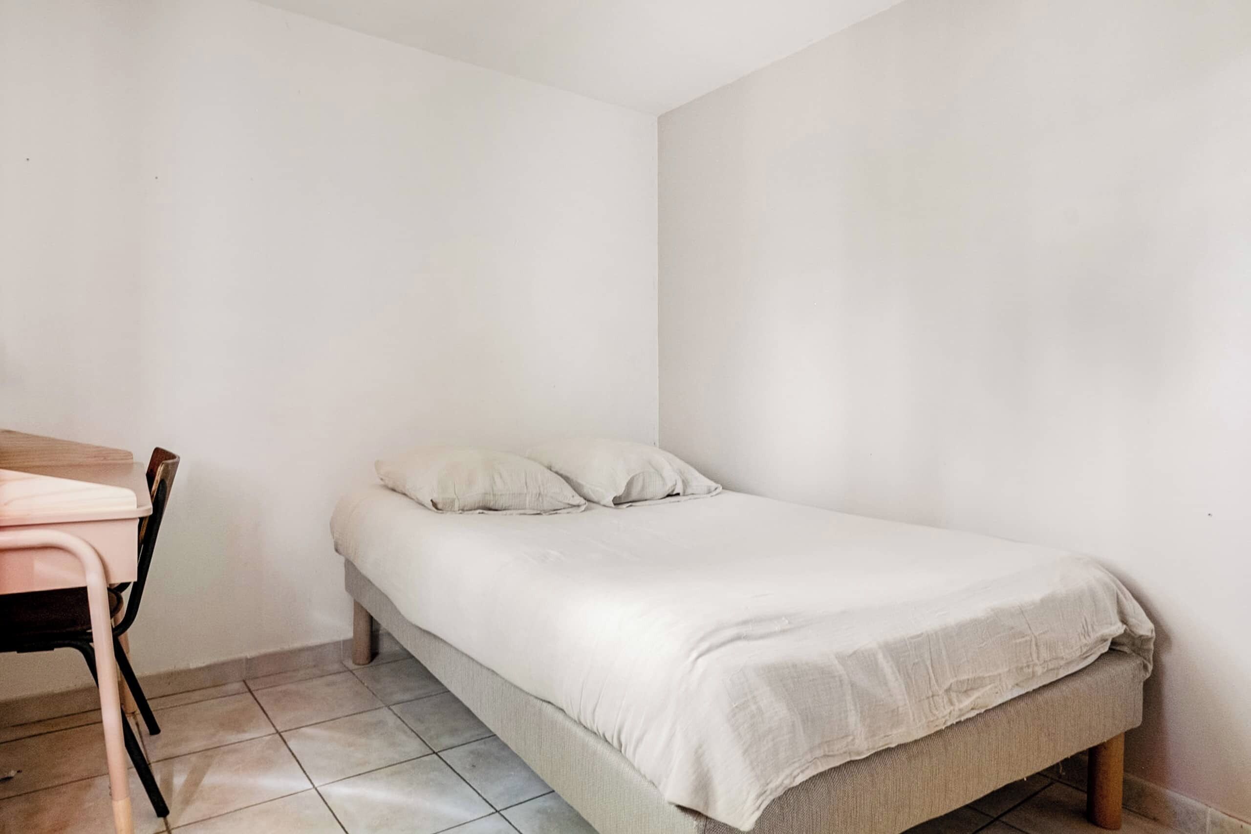 A small, single-bed room with a wooden desk and chair, plain white walls, and tiled flooring.