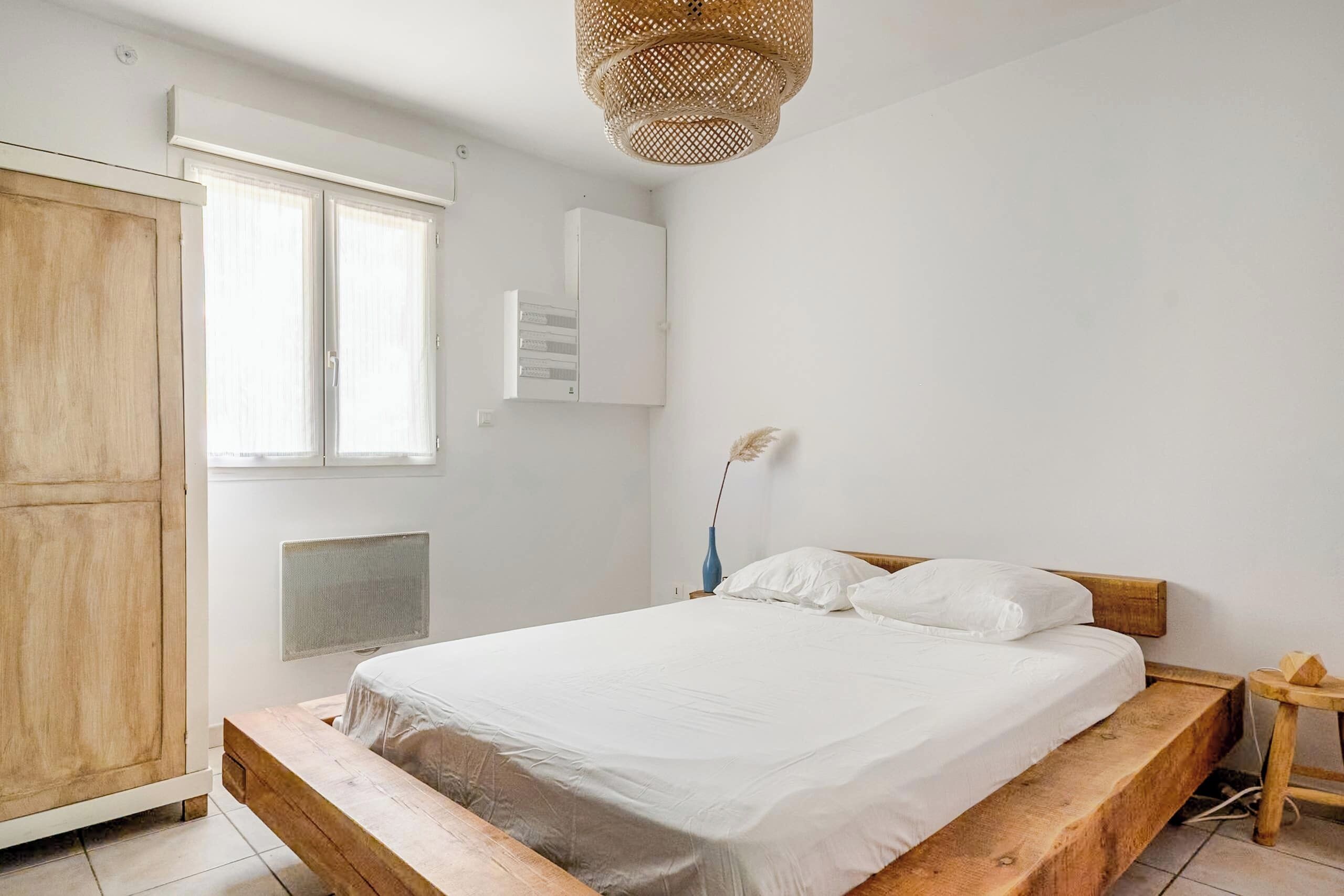 Ein Schlafzimmer mit hölzernem Bettgestell, weißer Bettwäsche, einem Kleiderschrank, einem Fenster und einer Wand-Klimaanlage.
