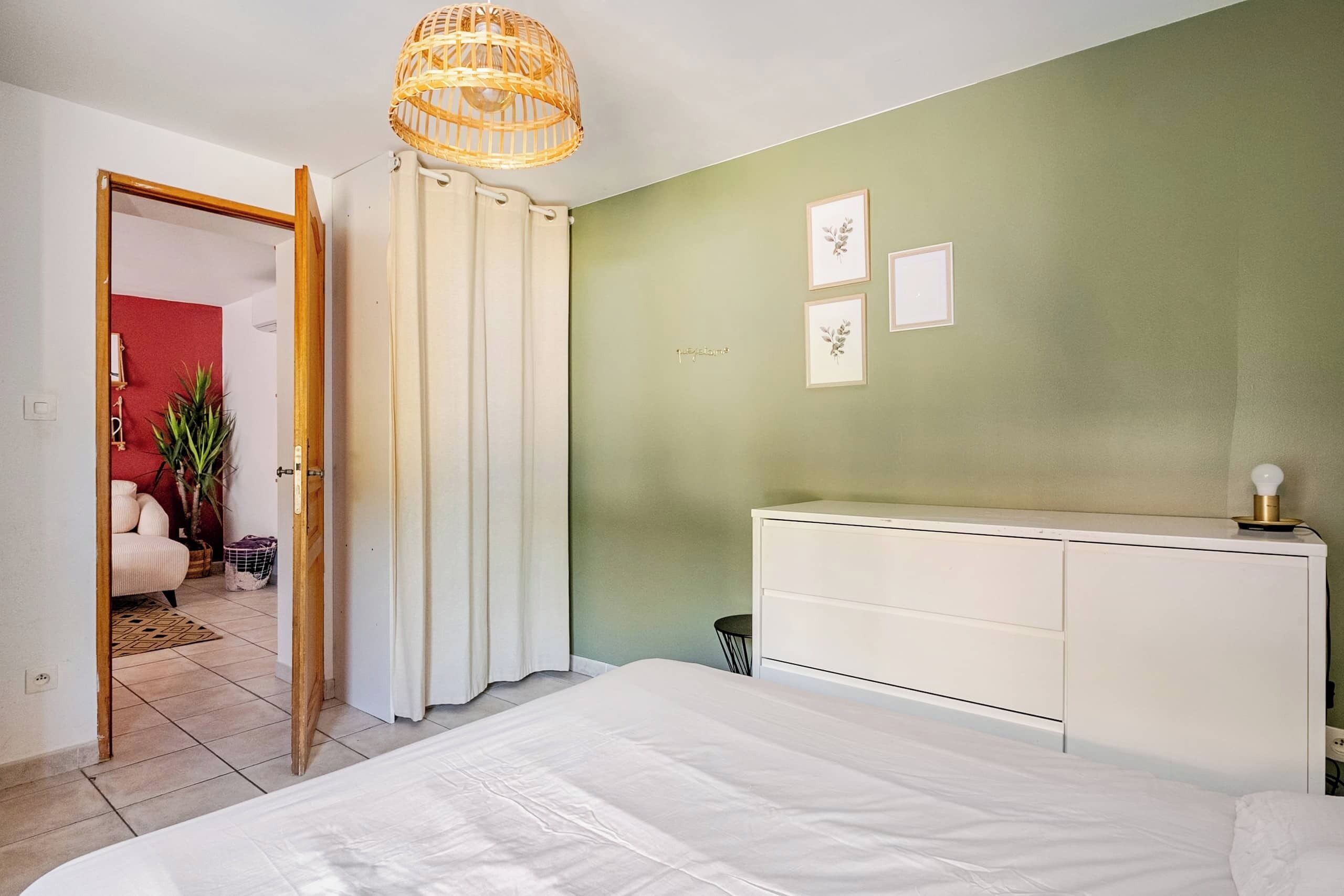 Une chambre à coucher comprenant un lit, une table de chevet, une armoire, un luminaire suspendu et une porte donnant sur une autre pièce.