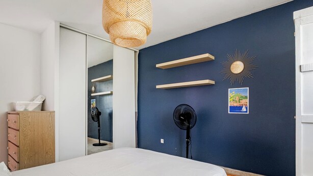 Un dormitorio con cama, cómoda, espejo, dos ventiladores y estantes en la pared.