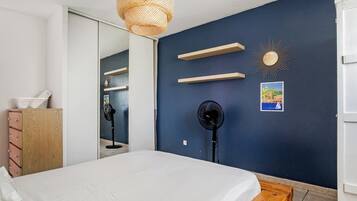 Een slaapkamer met een bed, een dressoir, een spiegel, twee ventilatoren en wandplanken.