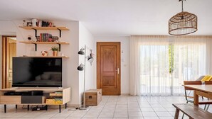 Una sala de estar moderna con televisor de pantalla plana, estanterías de madera, una mesa de comedor y un amplio ventanal con cortinas translúcidas.
