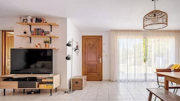 Een moderne woonkamer met een flatscreen televisie, houten planken, een eettafel en een groot raam met dunne gordijnen.