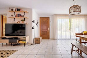 Ein modernes Wohnzimmer mit Flachbildfernseher, Holzregalen, einem Esstisch und einem großen Fenster mit durchscheinenden Vorhängen.