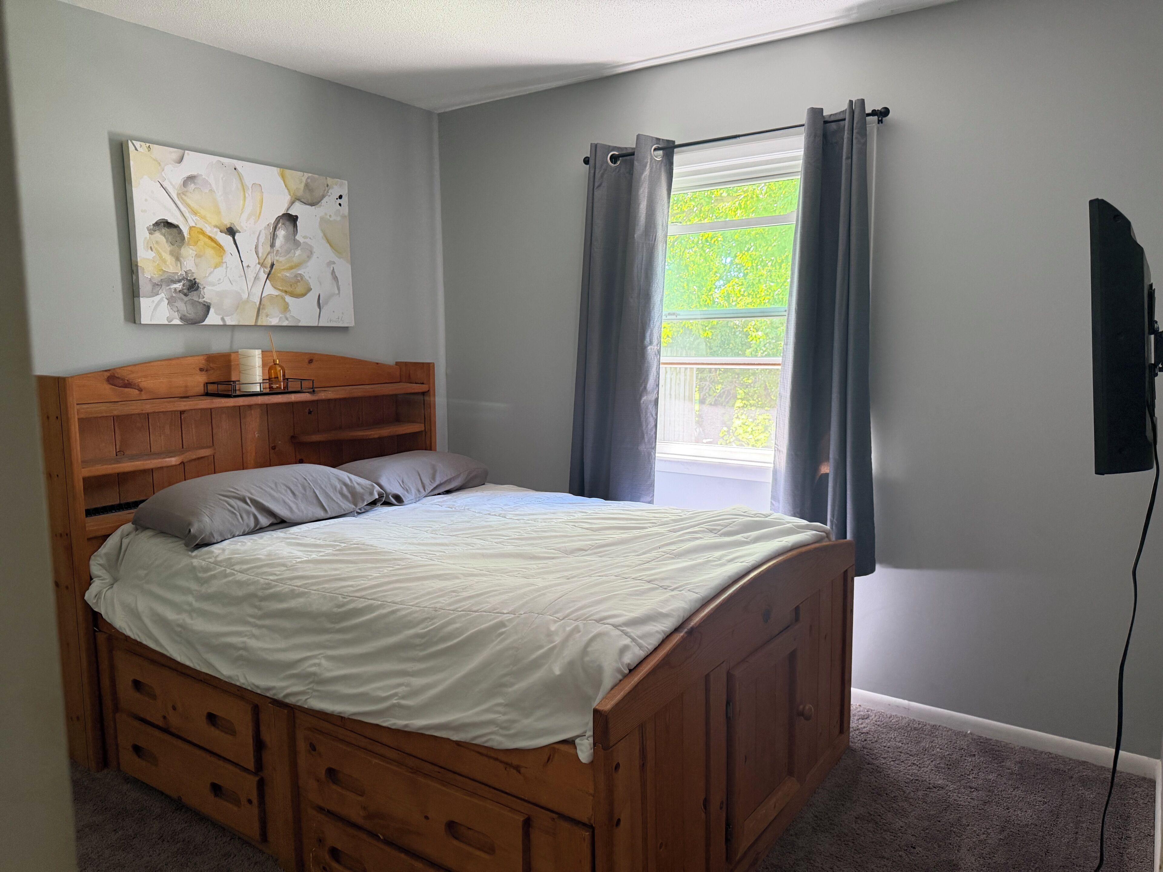 A bedroom with a wooden bed, a nightstand, a wall-mounted TV, a window with curtains, and a painting on the wall.