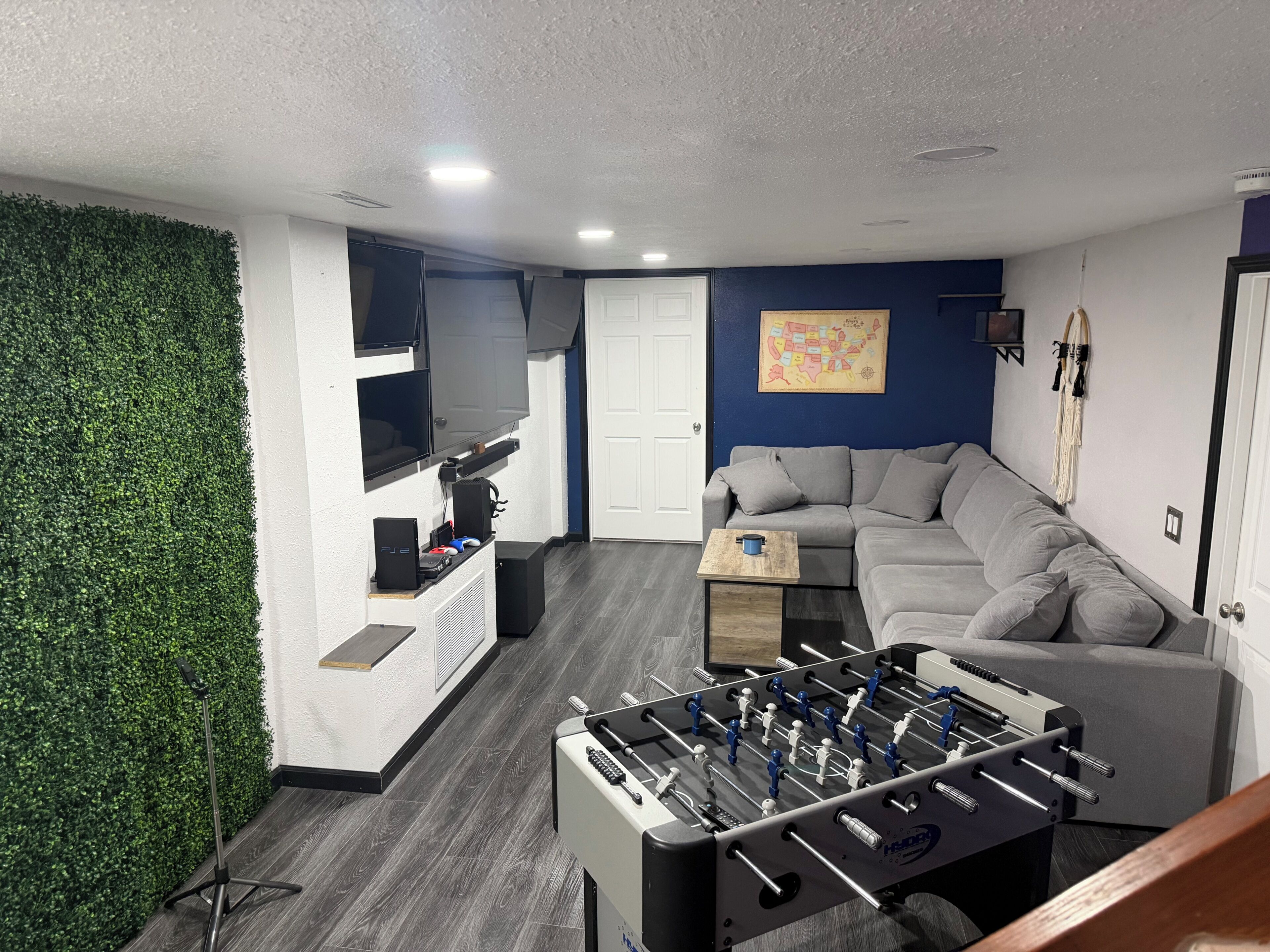 A room with a foosball table, a gray sofa, a TV mounted on the wall, and a green wall.