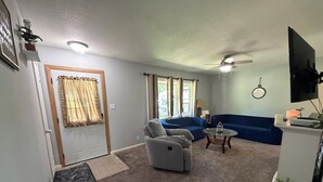 A living room with a sofa, armchair, and ottoman, a television, and a ceiling fan. - 4-BR2-BA House INSIDE Iowa State fair! With jetted tub for 2 (Des Moines)