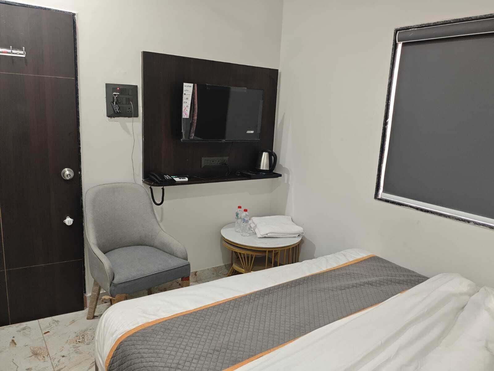 A hotel room with a bed, a chair, a small table with a towel and water bottles, a TV mounted on the wall, and a door.