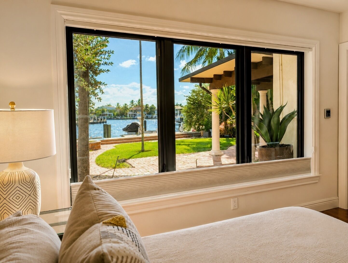 A room with a large window, a bed, a lamp, and a view of a landscaped area with a pool and palm trees.