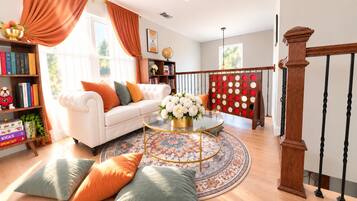 A living room with a white sofa, a round coffee table, and a patterned rug. There is a bookshelf with books and decorative items, and a staircase with a wooden railing.