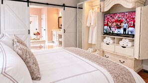 A bedroom with a large bed, a TV displaying a football game, and a walk-in closet with towels and robes.