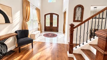 A spacious living room with a black sofa, wooden flooring, a staircase with a wooden railing, and a large wooden door with decorative glass panels.