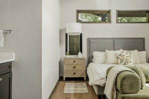 A bedroom with a bed, bedside table, lamp, and a view of greenery through windows. - Charming Lakeside Cabin | BBQ Grill | Pickleball (Lake Travis)