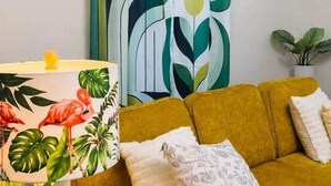A room with a yellow sofa, a patterned lamp, a green and white abstract curtain, and wall-mounted shelves with plants.